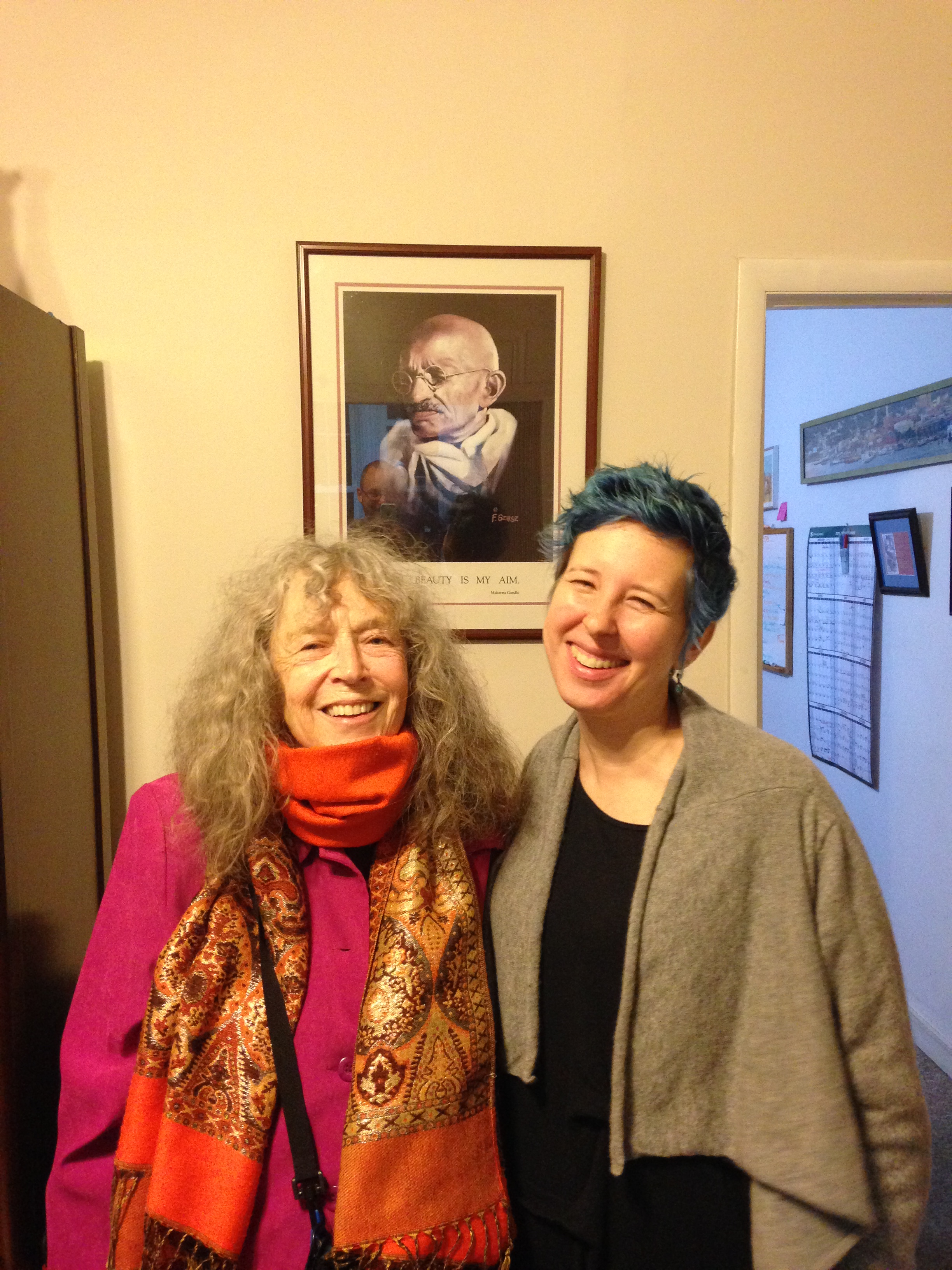 Two people smiling in a room, one with long curly hair in a pink coat and colorful scarf, the other with short blue hair wearing a dark shirt and gray cardigan. A framed portrait hangs on the wall behind them.