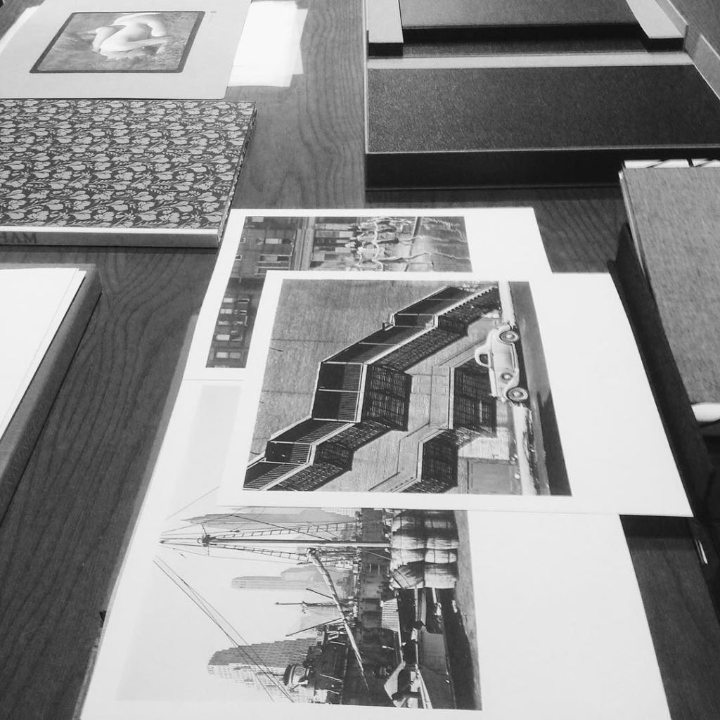 Black and white photo showing several prints and books on a table. Images include a car near a geometric building, people in a park, and a harbor scene with ships. Some books are open, displaying patterned covers.