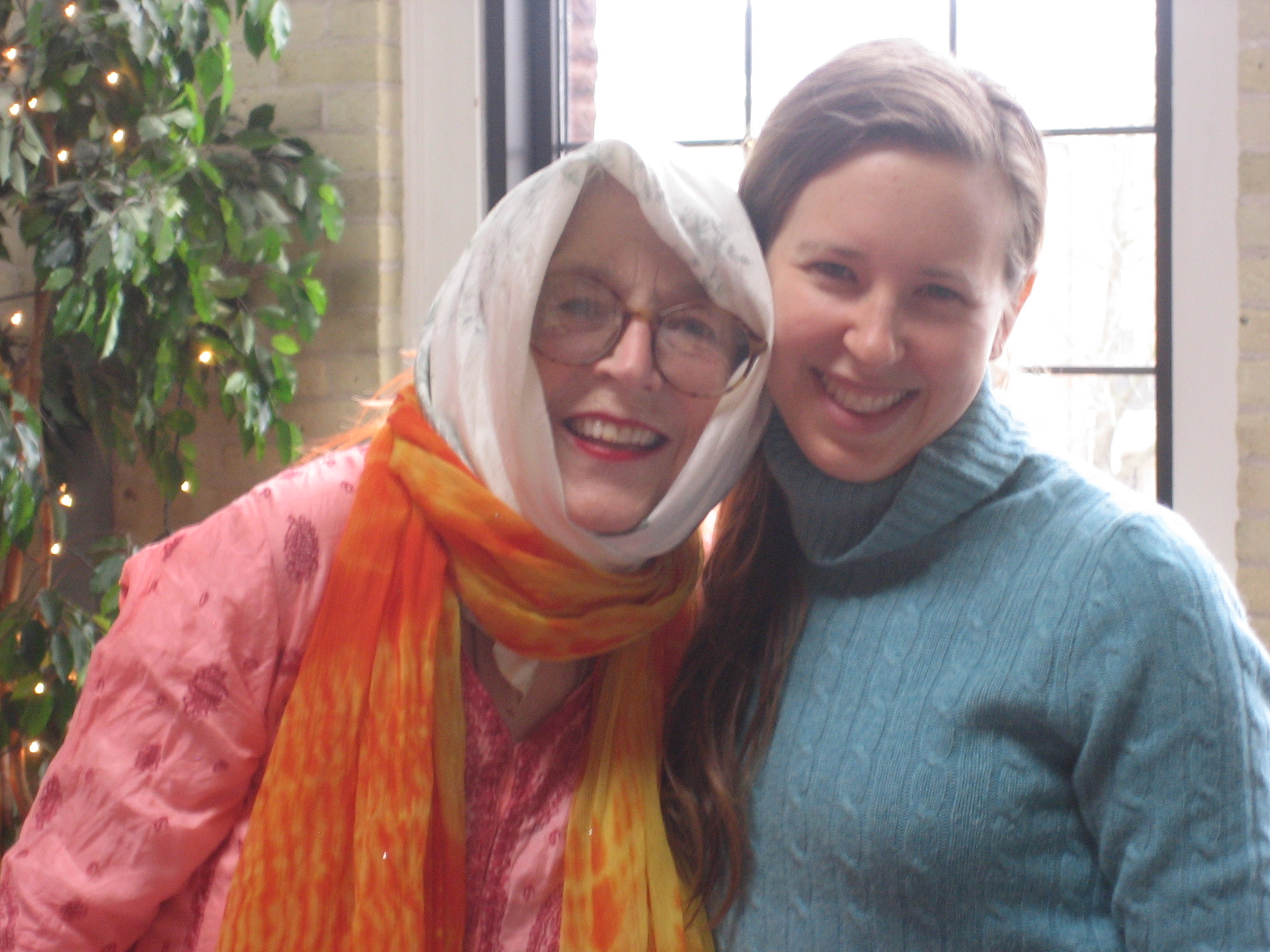 Two women smiling indoors. The older woman wears glasses, a colorful scarf, and a pink outfit. Karen Gonzalez Rice, with long hair, wears a blue sweater. They stand close together in front of a window and a potted plant.