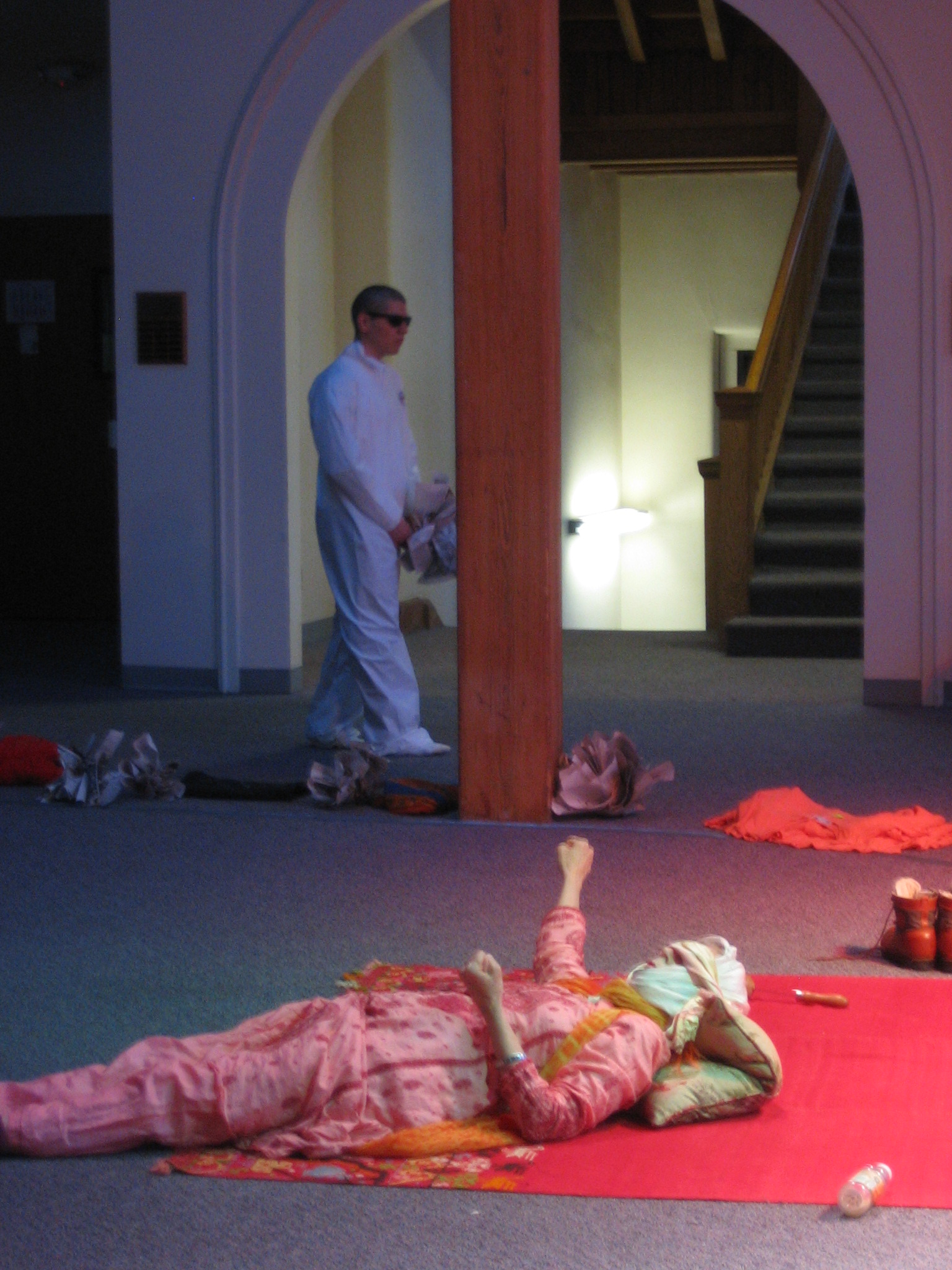 A person in a pink, patterned outfit lies on the floor with a bottle nearby. Another person in white clothing walks in the background, partially obscured by a wooden pillar. The setting appears to be indoors with a staircase visible.