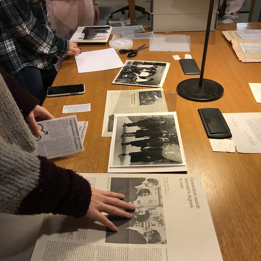 Students gathered around a wooden table examining black-and-white photographs and documents. Scissors, a phone, and a magnifying glass are visible, suggesting a focus on research or archival work.