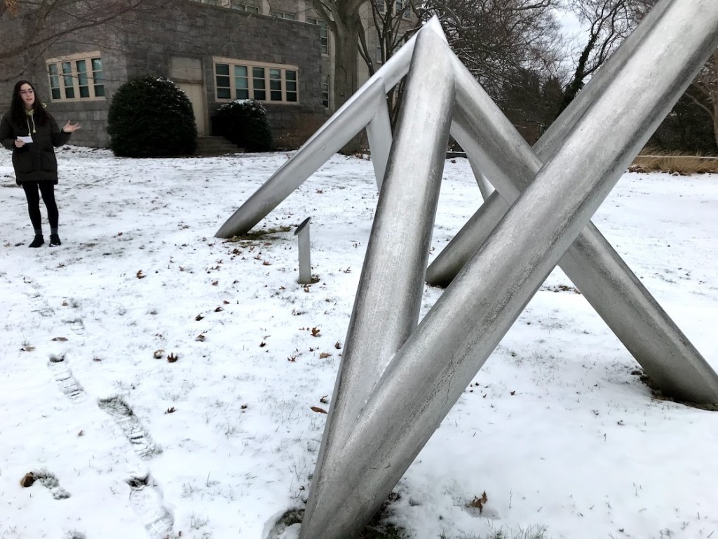 A snow-covered outdoor scene features a large, metal geometric sculpture composed of several angled beams. A person stands nearby, dressed in winter clothing, with tracks in the snow leading towards the sculpture.