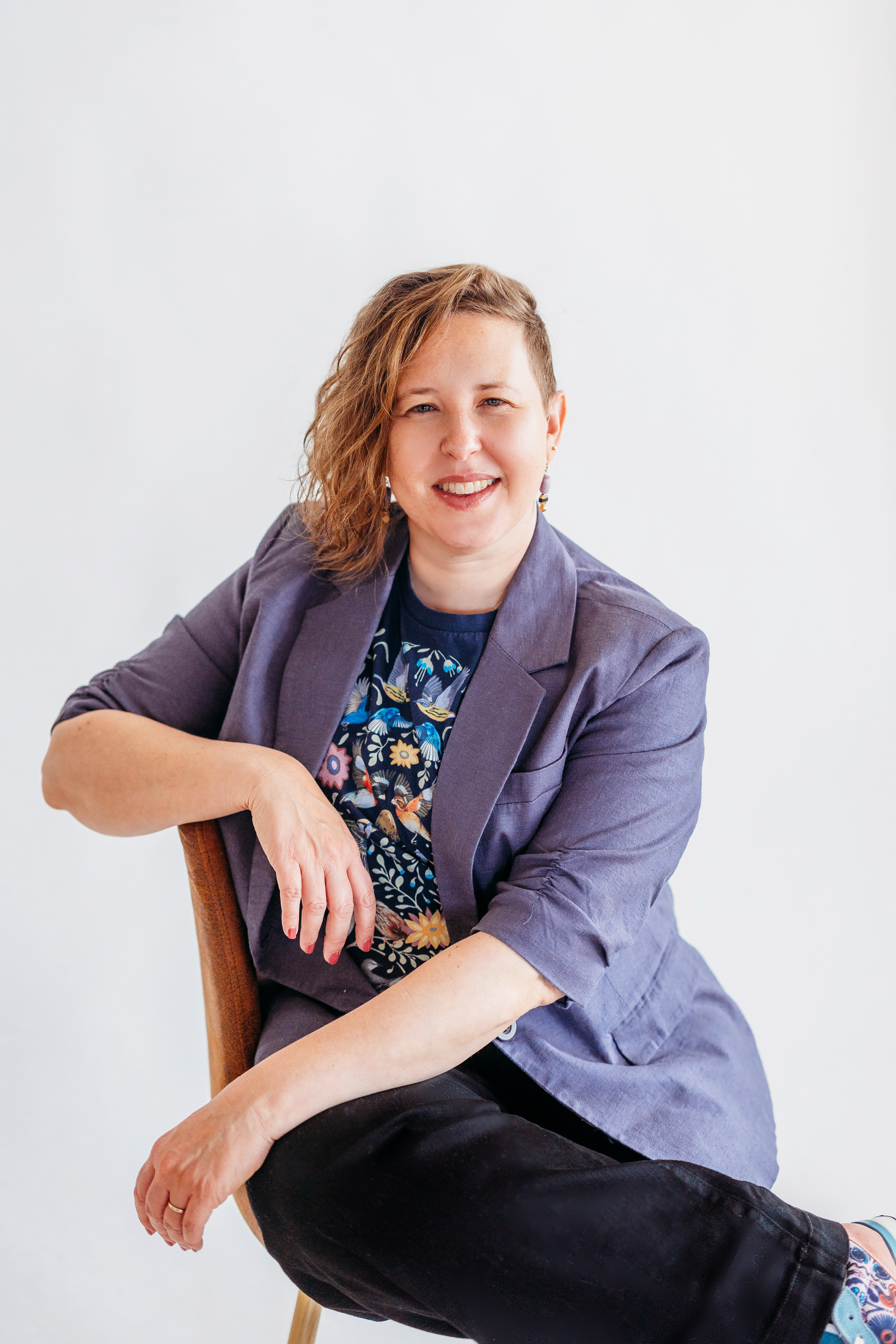 Karen Gonzalez Rice with short, wavy hair smiles while seated on a wooden chair. They wear a gray blazer over a floral shirt and black pants. The background is a plain, light color.