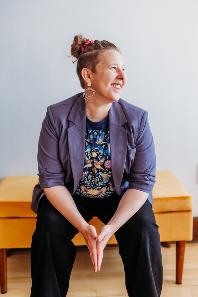 Karen Gonzalez Rice with short hair sits smiling on a yellow bench, wearing a blue blazer, a patterned shirt, and black pants. They have large hoop earrings and a bun tied with a pink scrunchie. The background is a plain light-colored wall.