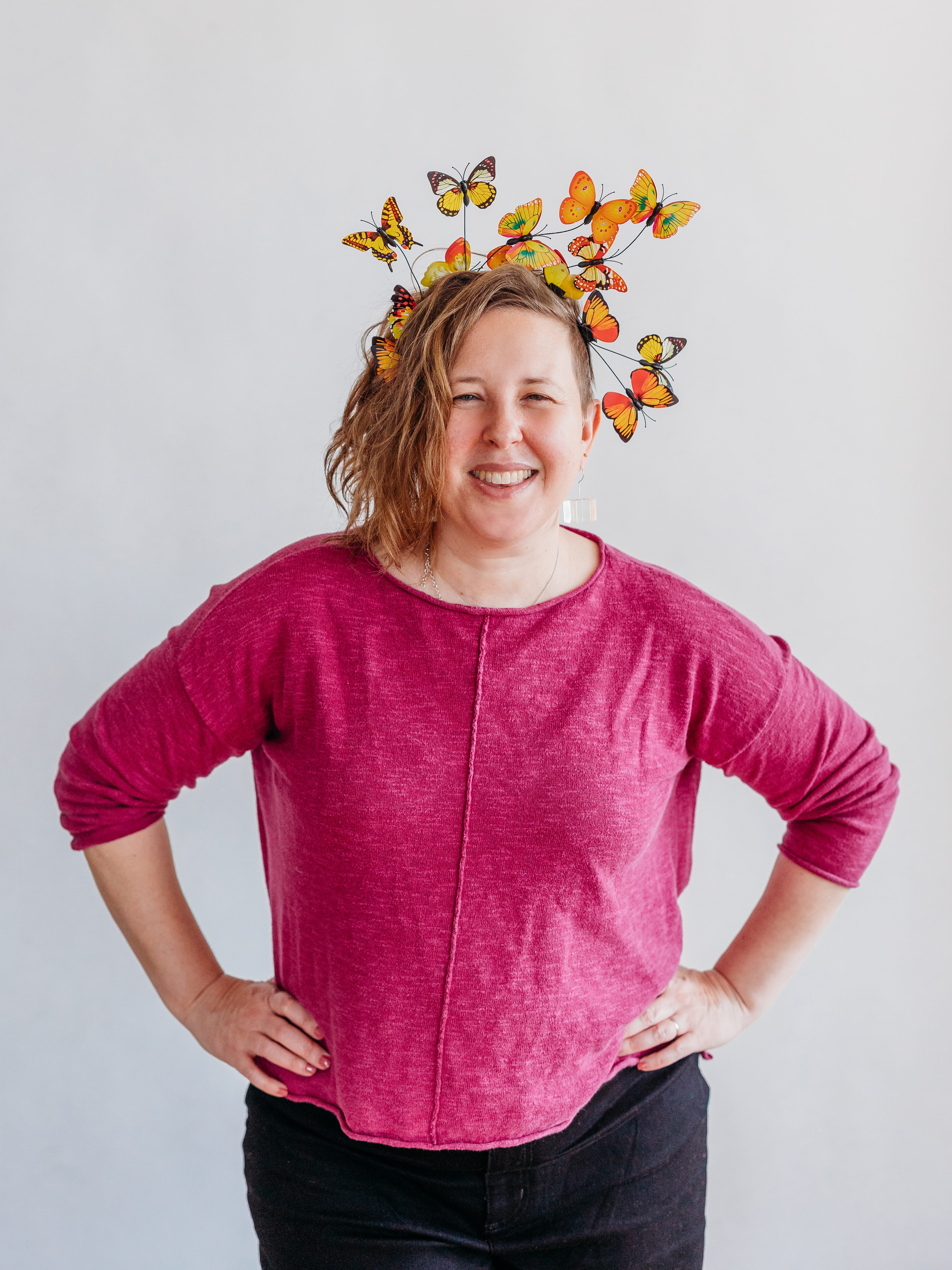 Karen Gonzalez Rice wearing a pink sweater stands against a white background, smiling with hands on hips. They are wearing a headband adorned with colorful butterflies resting on wires, creating a whimsical look.