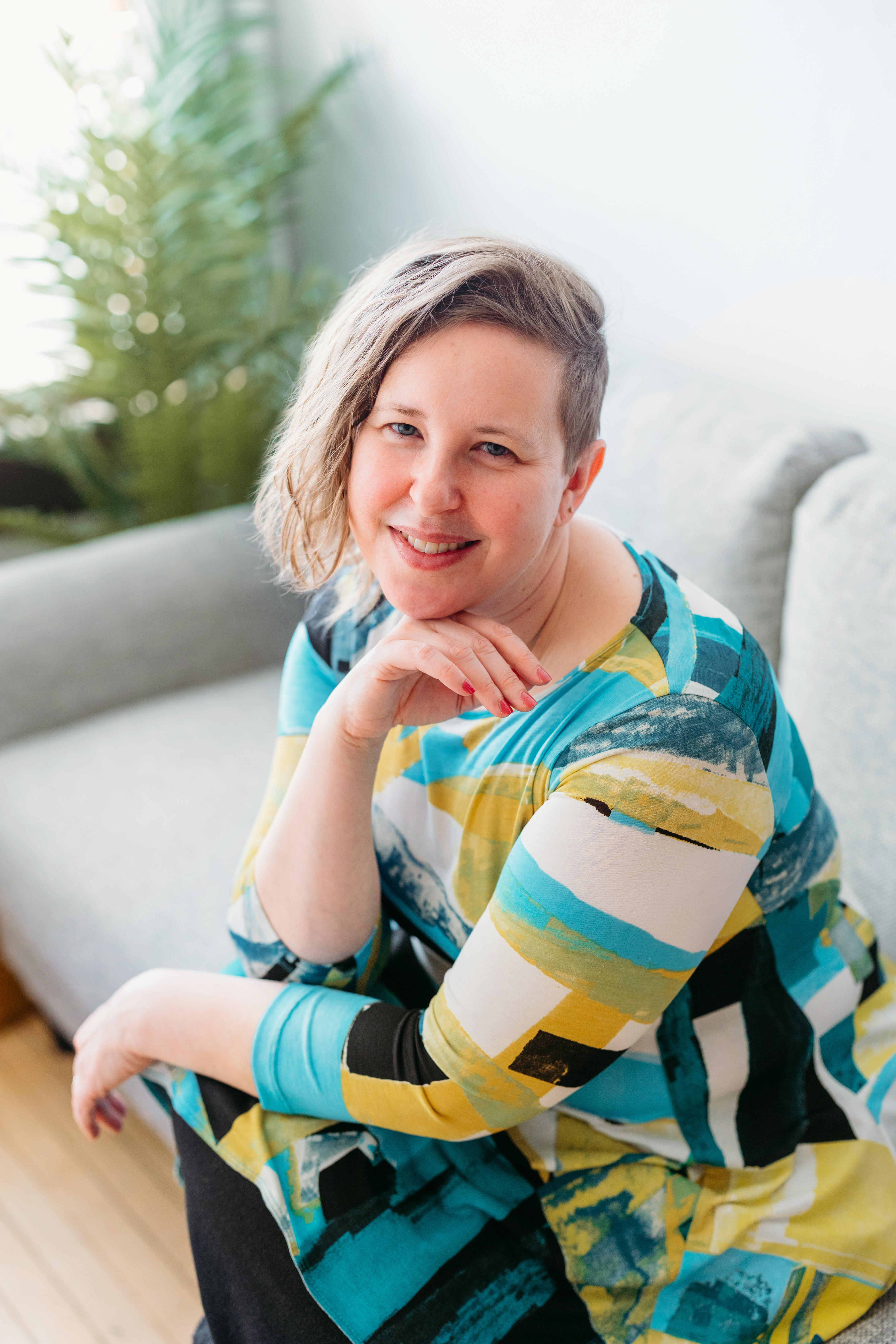 Karen Gonzalez Rice with short hair smiles warmly at the camera, sitting on a light gray couch. They are wearing a colorful, patterned dress with shades of turquoise, yellow, and black. A green plant is visible in the background.